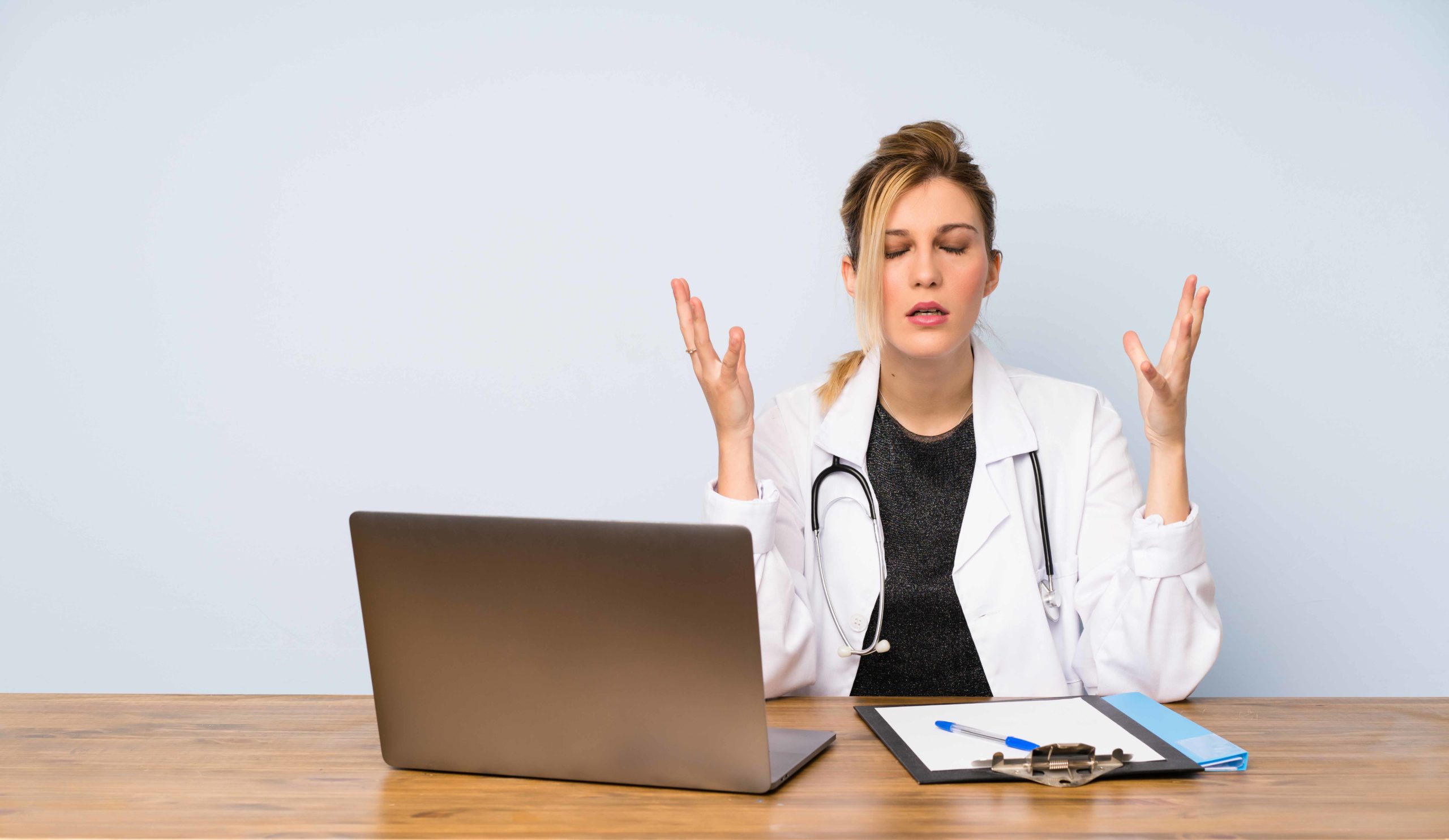 ノートPCとバインダーが置かれた机の前で、両手を挙げて険しい表情をしている女性医師